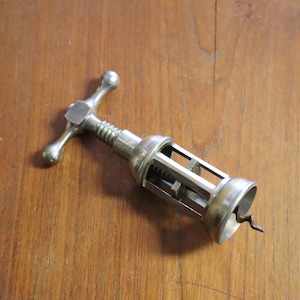 Può includere: Un cavatappi vintage color argento con manico e vite. Il cavatappi è appoggiato su una superficie di legno. Il design include un meccanismo a ingranaggi e una base a forma di campana.