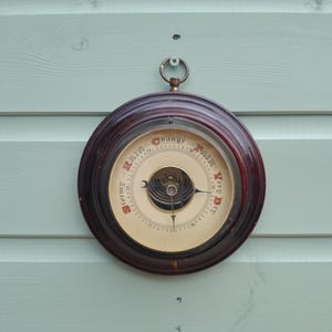 May include: A circular wall-mounted barometer with a dark brown wooden frame and a cream-colored dial. The face features black text reading Stormy, Rain, Change, Fair, and Very Dry, with a central brass mechanism and a single black metal pointer.