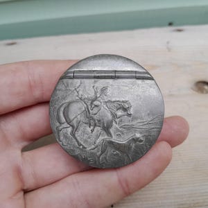 Puede incluir: Un estuche compacto redondo de metal plateado con una escena en relieve de una persona montando a caballo con un perro corriendo al lado. El recipiente circular tiene una tapa con bisagras en la parte superior con un diseño detallado.