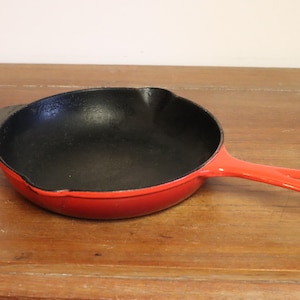 Könnte beinhalten: Eine rote gusseiserne Bratpfanne mit schwarzem Innenraum. Die Pfanne hat einen langen Griff und eine kleine Schlaufe auf der gegenüberliegenden Seite. Die Pfanne steht auf einer Holzoberfläche.