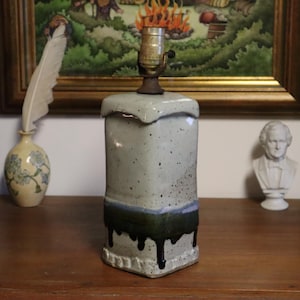 May include: A ceramic lamp with a square base and a light blue and green glaze. The lamp has a brass lamp holder and a unique, textured design. The base has a dark green drip effect. Decorative home decor.