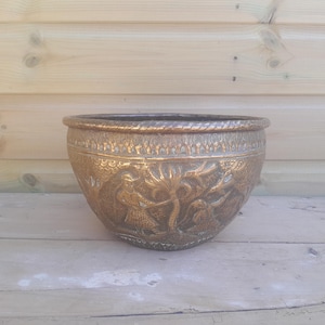 Pode incluir: Um vaso redondo de cor bronze com entalhes em relevo detalhados. O vaso tem uma borda larga com um design semelhante a uma corda e apresenta uma cena com figuras e folhagens. Ele está em uma superfície de madeira.