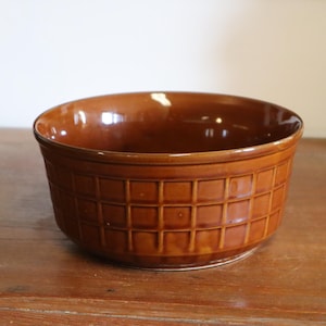 May include: A brown ceramic bowl with a grid pattern around the exterior. The bowl has a glossy finish and sits on a wooden surface. A decorative vase is visible in the background.