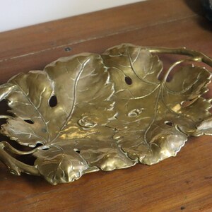May include: A brass leaf-shaped tray with intricate detailing. The tray features a central leaf design with smaller leaves and a curved handle. The brass has a warm, golden hue, and the tray is placed on a wooden surface.