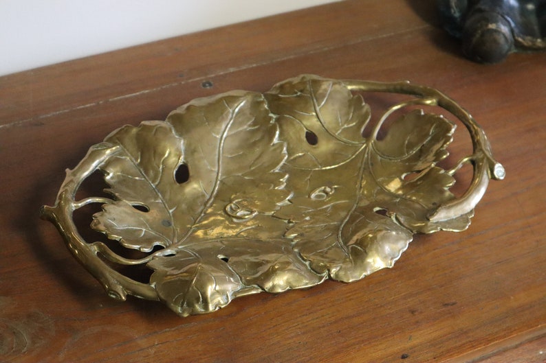 May include: A brass leaf-shaped tray with intricate detailing. The tray features a central leaf design with smaller leaves and a curved handle. The brass has a warm, golden hue, and the tray is placed on a wooden surface.