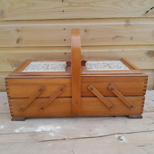 May include: A vintage wooden sewing box with a handle. The box is made of light brown wood and features a hinged lid with a floral pattern. It has two pull-out drawers and is designed for storing sewing supplies.