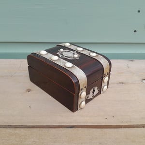 May include: A dark brown wooden box with a curved lid, featuring silver-toned metal bands secured by white circular studs. The box includes a central silver-toned decorative plate on the top and a matching silver-toned keyhole plate on the front.