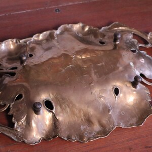 May include: A brass-colored decorative tray in a leaf design. The tray has a central leaf-shaped area with several small holes and is surrounded by a decorative border. The tray has four small, dark-colored knobs.