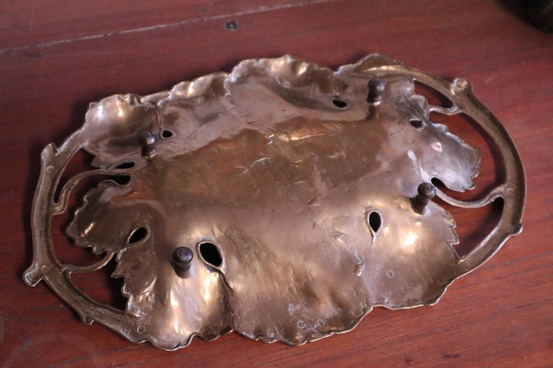May include: A brass-colored decorative tray in a leaf design. The tray has a central leaf-shaped area with several small holes and is surrounded by a decorative border. The tray has four small, dark-colored knobs.