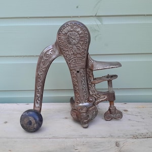 Könnte beinhalten: Ein antiker Apfelschäler aus Gusseisen mit verziertem Reliefdesign, ausgestattet mit einem geschwungenen Griff mit dunklem, kugelförmigem Knauf, einem mechanischen Getriebegehäuse mit der Aufschrift ORIGINAL SAFETY und einer Schraubzwinge.