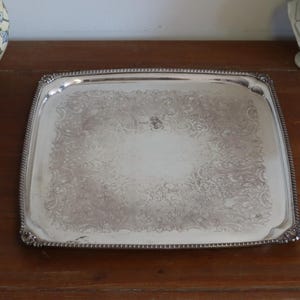 Könnte beinhalten: Ein rechteckiges silbernes Serviertablett mit kunstvollen floralen Mustern. Das Tablett hat einen Perlenrand und abgerundete Ecken. Es wird auf einer Holzoberfläche präsentiert, mit dekorativen Gegenständen im Hintergrund.
