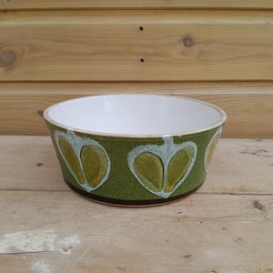 May include: A round ceramic bowl with a white interior and a textured olive green exterior. The outer surface features a repeating pattern of stylised, light green heart-shaped fruit motifs outlined in white, with a dark brown band at the base.