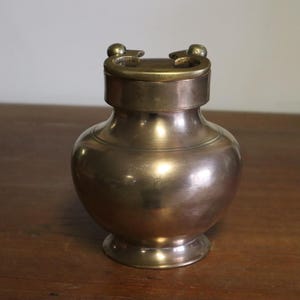 May include: A vintage brass vase with a rounded body and a lid with two small handles. The vase has a polished finish and sits on a wooden surface. The design is simple and elegant, suitable for home decor.