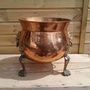 May include: A large, hammered copper cauldron with a wide rim and ornate lion's head handles. The cauldron rests on three decorative, claw-footed legs. The copper has a rich, warm colour, and the cauldron is likely used for decorative purposes.