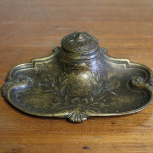 May include: Antique brass inkwell with a decorative floral design. The inkwell features a central ink pot with a lid, set on a tray with ornate edges. The brass has a weathered, aged appearance, suggesting its vintage origin.