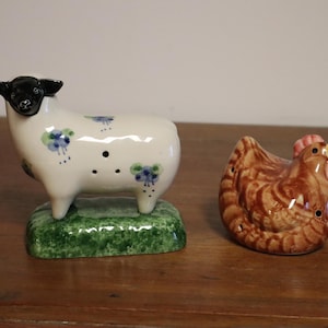 May include: A ceramic sheep and chicken figurine set. The sheep is white with black ears and a black face, and is decorated with blue floral accents. The chicken is brown with a red comb and wattle. Both are likely salt and pepper shakers.