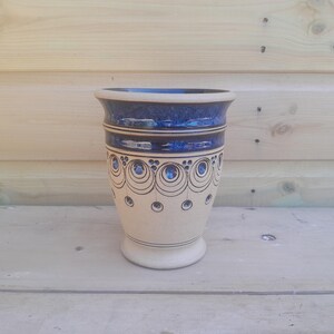 Pode incluir: Um vaso de cerâmica com base creme e borda azul escuro. O vaso apresenta um padrão decorativo de círculos azuis e linhas pretas. O vaso tem aproximadamente 20 cm de altura.