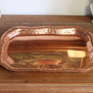 May include: A rectangular copper tray with a hammered texture and a slightly irregular edge. The tray reflects the surrounding environment, showcasing a warm, metallic sheen. The tray is displayed on a wooden surface.