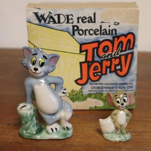 May include: Two Wade porcelain figurines of Tom and Jerry, with a box behind them. Tom, a grey cat, leans on a green base, and Jerry is a small mouse. The box reads "WADE real Porcelain Tom and Jerry."