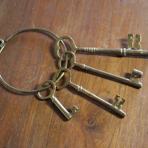 May include: A set of antique brass keys on a ring, resting on a wooden surface. The keys have ornate designs and varying shapes. The ring is circular with a small loop. The keys are arranged in a cluster, suggesting a vintage or decorative theme.