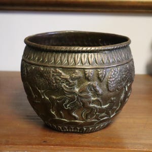 May include: A bronze-coloured, round decorative bowl with detailed relief carvings of figures on horseback and foliage. The rim has a decorative pattern. A blue vase and a white bust are in the background.