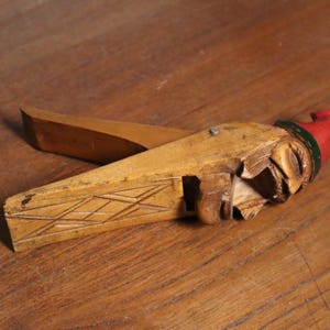 May include: A vintage wooden nutcracker in the shape of a figure with a red hat and green trim. The nutcracker is carved from light-colored wood and features a lever for cracking nuts. The design includes etched details.