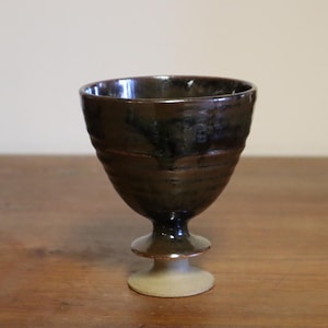 Op de afbeelding: Een handgemaakte keramische beker met een donkerbruin glazuur. De beker heeft een brede, getextureerde kom en een voet. De beker is ongeveer 13 cm hoog en wordt op een houten oppervlak getoond.