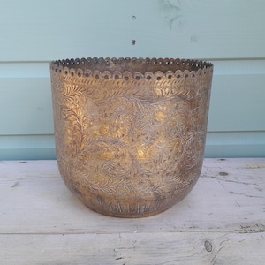 Pode incluir: Um vaso cilíndrico de latão com um intrincado padrão floral gravado na sua superfície exterior. O rebordo superior apresenta um acabamento decorativo recortado e o metal exibe uma pátina envelhecida com tons escuros.