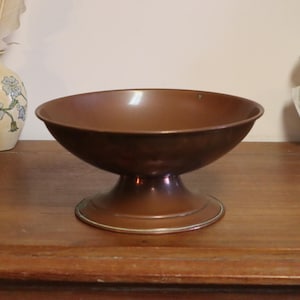 May include: A copper-coloured pedestal bowl with a wide, shallow basin. The bowl sits on a rounded base with a silver-coloured trim. A decorative vase and a bust are in the background.