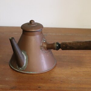 May include: An antique copper teapot with a wooden handle and lid. The teapot features a spout and decorative detailing. The handle is approximately 15 cm long. The teapot is displayed on a wooden surface.