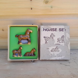 May include: A vintage "HORSE SET" box with three brown ceramic horse figurines. The box is open, revealing the figurines on a green background. The box cover has illustrations of horses.