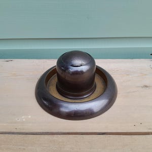 May include: A dark brown, circular inkwell featuring a central domed reservoir with a hinged lid, set within a wide, flat, matching circular base. The object has a smooth, polished finish and a deep, uniform colour throughout its structure.