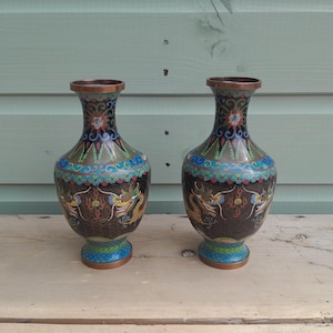 May include: A pair of matching cloisonné vases featuring a dark brown background with intricate multi-coloured dragon motifs, floral patterns, and geometric borders. The vases have a metallic rim and base, showcasing detailed enamel work in shades of blue, green, yellow, and red across the surface.