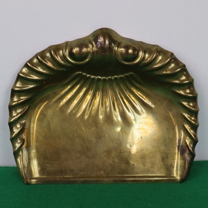 May include: A vintage brass dustpan with a shell-like design. The dustpan has a wide, curved rim with a decorative edge and a rectangular base. The brass has a warm, golden hue, and the overall design is ornate and elegant.
