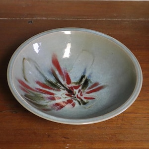 May include: A handmade ceramic bowl with a light blue glaze. The interior features a floral design in red, green, and black. The bowl is round and sits on a wooden surface.