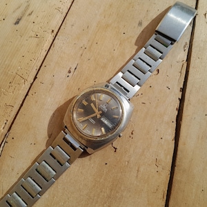 Puede incluir: Reloj de pulsera Titan vintage con una correa de metal plateado y una esfera dorada. El reloj tiene una esfera gris oscuro con manecillas e indicadores dorados. Se muestra sobre una superficie de madera envejecida.