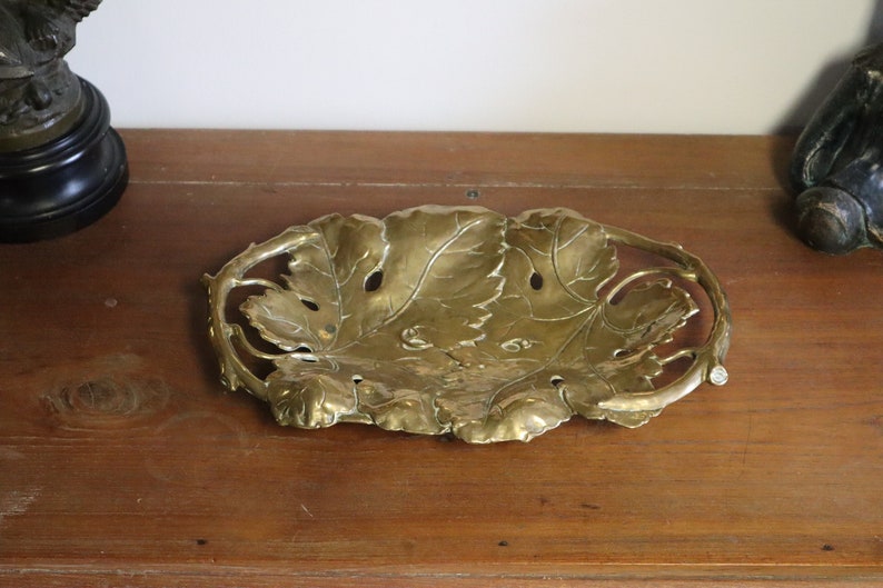 May include: A decorative brass leaf-shaped tray. The tray features a detailed design of leaves and stems, with a slightly raised edge. The brass has a warm, golden hue, and the tray is placed on a wooden surface.