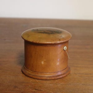 May include: A small, antique wooden box with a rounded lid and base. The box is cylindrical with a light brown finish and a small, white loop on the side. The lid has a darker, possibly painted, design.