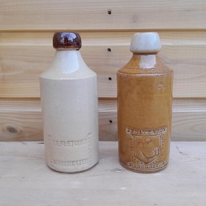 May include: Two vintage stoneware bottles with cylindrical bodies and narrow necks. The left bottle is cream-colored with a dark brown cap and text reading T.E. FLETCHER MANCHESTER. The right bottle is tan with a light cap and text reading J. PRATT & SON MANCHESTER.
