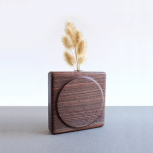May include: A dark brown wooden vase with a circular opening, featuring a simple, modern design. The vase is filled with dried, light brown grasses.