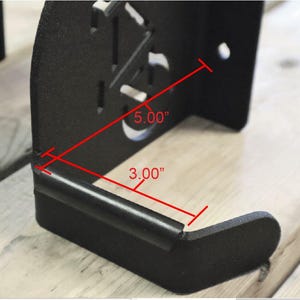 Puede incluir: Soporte de pared de metal negro con forma rectangular de 5,00" x 3,00". El soporte tiene un borde redondeado y una peque&ntilde;a barra de metal que se extiende desde la parte inferior.