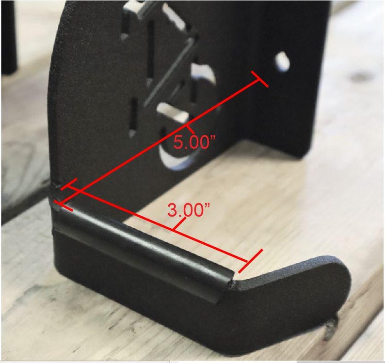 Puede incluir: Soporte de pared de metal negro con forma rectangular de 5,00" x 3,00". El soporte tiene un borde redondeado y una peque&ntilde;a barra de metal que se extiende desde la parte inferior.