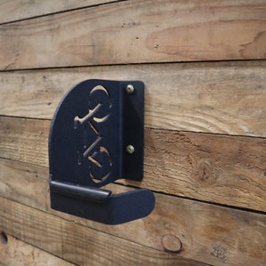 Puede incluir: Gancho para bicicleta de metal negro montado en una pared de madera. El gancho tiene forma de bicicleta con un dise&ntilde;o recortado.