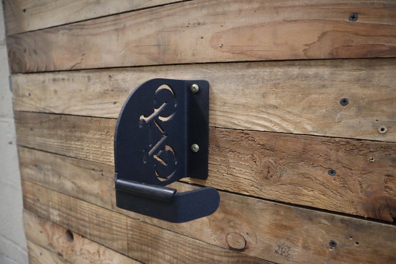 Puede incluir: Gancho para bicicleta de metal negro montado en una pared de madera. El gancho tiene forma de bicicleta con un dise&ntilde;o recortado.