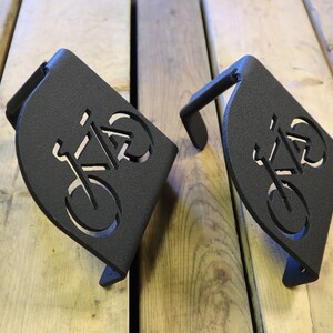 Puede incluir: Dos ganchos para bicicletas de metal negro con un dise&ntilde;o de bicicleta recortado. Los ganchos est&aacute;n montados en una superficie de madera.