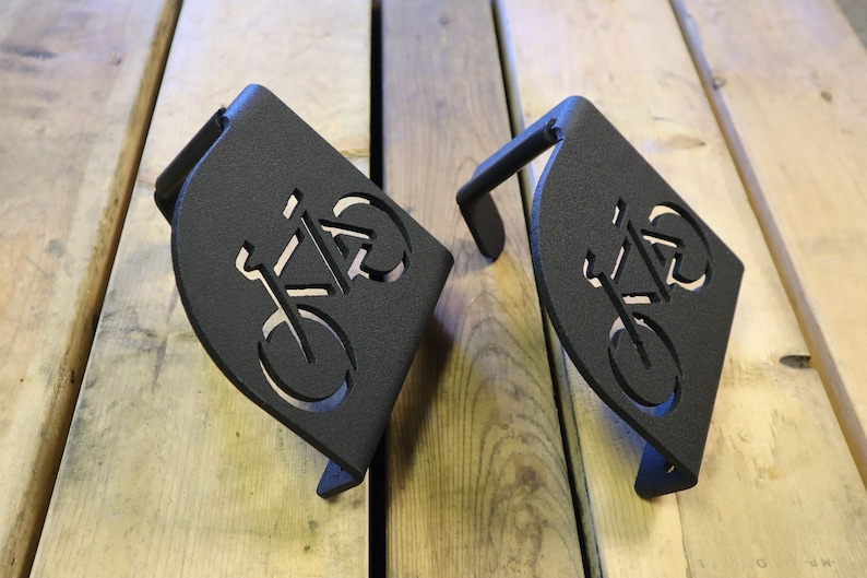 Puede incluir: Dos ganchos para bicicletas de metal negro con un dise&ntilde;o de bicicleta recortado. Los ganchos est&aacute;n montados en una superficie de madera.