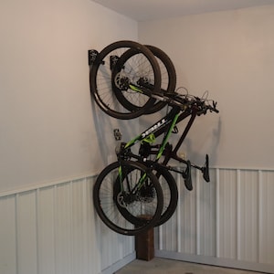 Puede incluir: Dos bicicletas de monta&ntilde;a negras est&aacute;n colgadas de un soporte de pared en un garaje. Las bicicletas est&aacute;n colgadas boca abajo con las ruedas mirando hacia la pared. Ambas bicicletas son negras con detalles verdes. La pared es blanca con un revestimiento de madera en la parte inferior.