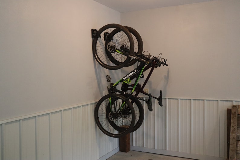 Puede incluir: Dos bicicletas de monta&ntilde;a negras est&aacute;n colgadas de un soporte de pared en un garaje. Las bicicletas est&aacute;n colgadas boca abajo con las ruedas mirando hacia la pared. Ambas bicicletas son negras con detalles verdes. La pared es blanca con un revestimiento de madera en la parte inferior.