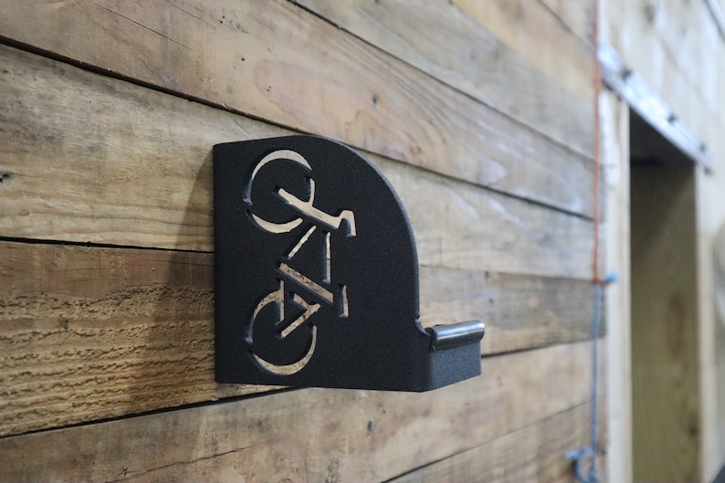Puede incluir: Portabicicletas de metal negro montado en una pared de madera. El soporte tiene forma de bicicleta con detalles recortados.