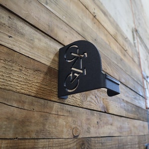 Puede incluir: Portabicicletas de metal negro montado en una pared de madera. El soporte tiene forma de bicicleta con un dise&ntilde;o recortado.